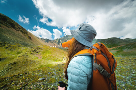 Successful Tourist Drinks Water In A Green Meadow Near High Mountains. Travel And Hiking Concept With Backpack, Side View