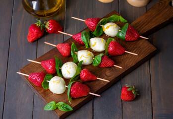 Strawberry Caprese Skewers. Lemon olive oil marinated mozzarella mini balls with fresh basil leafs and strawberries threaded on skewers served on a wooden board. Selective focus, horizontal.