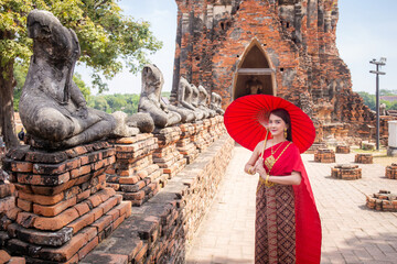 Young woman wearing traditional red Thai dress and golden accessories stands holding a traditional umbrella in the historical site at Wat Chaiwatthanaram, Ayutthaya, Thailand