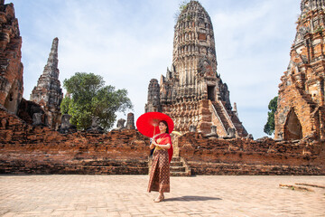 Young woman wearing traditional red Thai dress and golden accessories stands holding a traditional umbrella in the historical site at Wat Chaiwatthanaram, Ayutthaya, Thailand - obrazy, fototapety, plakaty