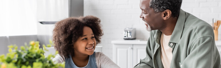 Fototapeta premium happy african american grandfather looking at curly granddaughter in kitchen, banner.