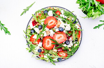 Strawberry and herbs healthy salad with arugula, blueberries, soft white feta cheese and walnuts, white kitchen table, place for text. Fresh useful dish for healthy eating