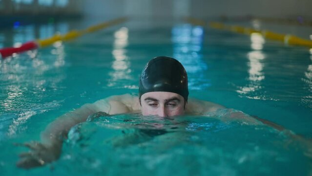 Front view male swimmer training movement in blue water in slow motion. Portrait of young motivated Caucasian man in cap swimming exercising in pool indoors