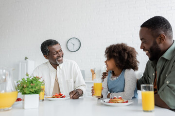 Fototapeta na wymiar happy middle aged man looking at african american granddaughter near son during family breakfast.