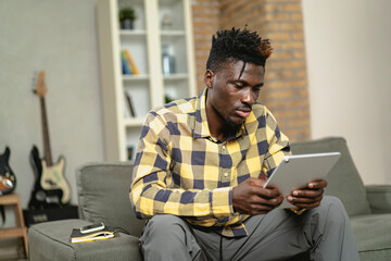 Young African man using digital tablet at home. Smiling man reading news while sitting on the couch at home.