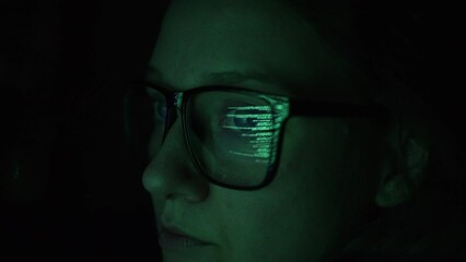 woman hacker programmer analyzes data in the dark close-up, on glasses is a green reflection of the program code from the screen of a laptop or computer, hacking the system and data security