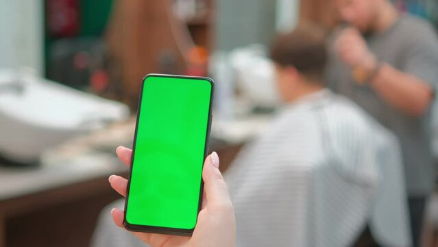 Close up Male Client watching at the smartphone green screen mockup while barber cuts his hair. Razors machine, tools, nozzles, and a hair dryer are visible in the background. Indoor business
