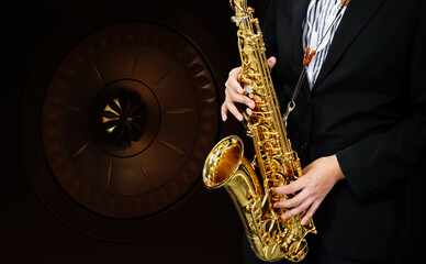 Male Musician in a Formal Black Suit Holds a Tenor Saxophone on Loudspeaker on dark background....