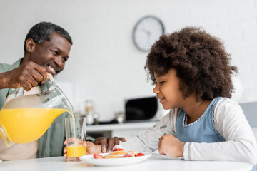happy african american girl looking at glass while grandfather pouring orange juice during breakfast.