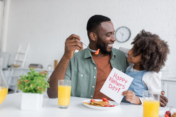 cheerful african american girl holding greeting card with happy fathers day lettering near smiling dad during breakfast.