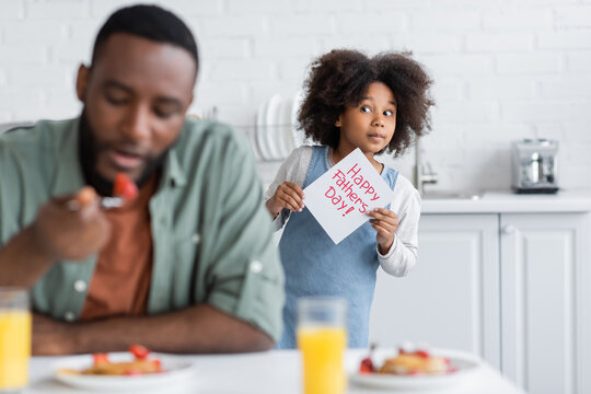african american girl holding greeting card with happy fathers day lettering while standing behind dad on blurred foreground.