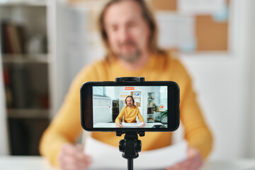 Mature teacher using mobile phone for recording online lesson, he sitting at table in class and...