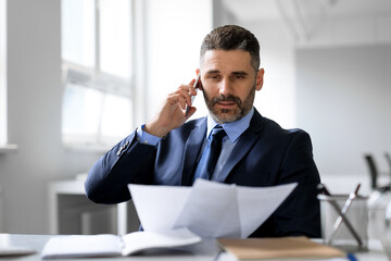 Naklejka premium Busy middle aged businessman talking on cellphone and checking papers, working with documents in office