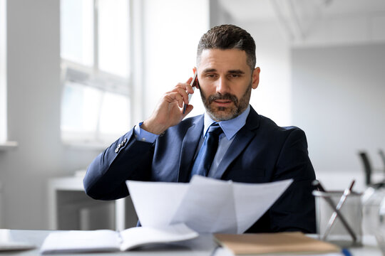 Busy middle aged businessman talking on cellphone and checking papers, working with documents in office