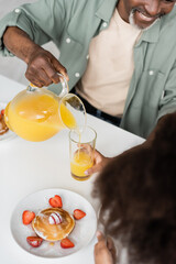 top view of happy african american man pouring orange juice near curly granddaughter during breakfast.