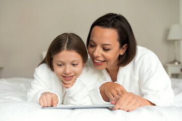 Cheerful european millennial woman and little girl in bathrobes watch video on tablet, lie on soft bed