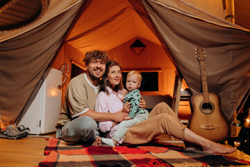 Happy family with lovely baby playing and spend time together in glamping on summer evening. Luxury camping tent for outdoor recreation and recreation. Lifestyle concept