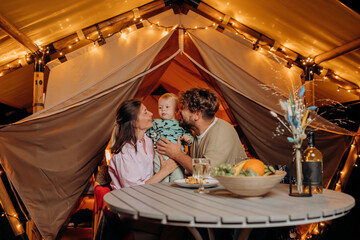 Obraz na płótnie Canvas Happy family with lovely baby have dinner and spend time together in glamping on summer evening near cozy bonfire. Luxury camping tent for outdoor recreation and recreation. Lifestyle concept