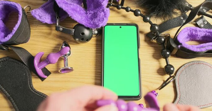 Man's hand examines a purple anal stimulator against the background of lying smartphone with green screen. Finger touches the application screen. Close-up.