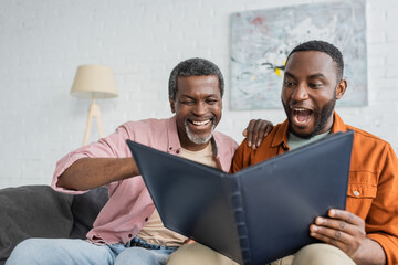 Cheerful and excited african american father and son looking at photo album at home.