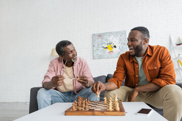 Overjoyed african american man playing chess with mature dad near smartphone with blank screen at home.