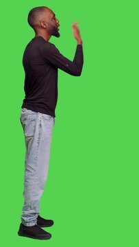Vertical video: Profile of african american man sending air kisses on camera, posing with kissy face on full body greenscreen. Young adult being romantic in studio and doing flirty gesture, optimistic