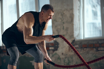 Obraz premium Coach. Young man with sportive, strong, muscular body keeping body toned, training with sports equipment, pulling battle ropes indoors. Concept of sportive lifestyle, body care, fitness, hobby, health