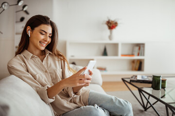 Glad millennial mixed race woman in wireless headphones look at phone, read message, chatting