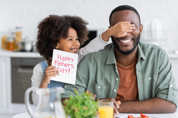 curly african american girl holding greeting card with happy fathers day lettering while covering eyes of dad.