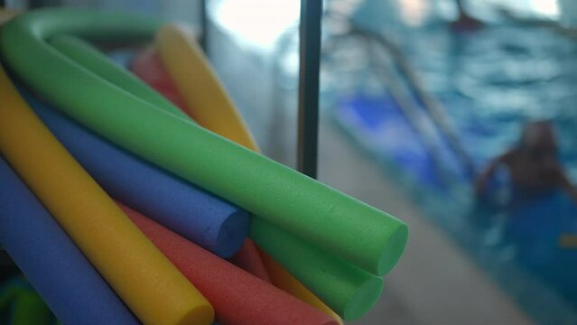Close-up colorful swimming sticks at poolside with blurred children training in water at background. Floatable toys in swimming pool indoors. Slow motion