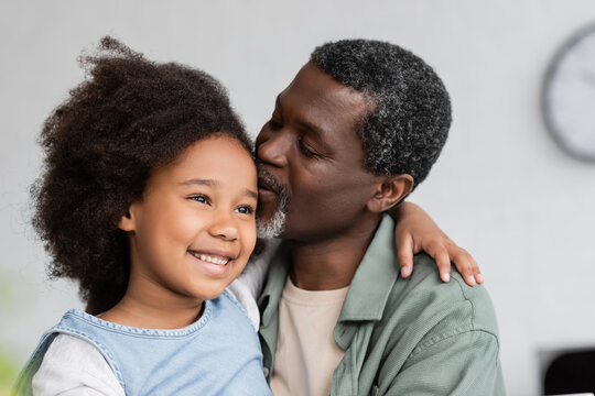 african american grandfather kissing head of cheerful granddaughter with curly hair.
