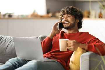 Young Indian Man Talking On Mobile Phone And Using Laptop At Home