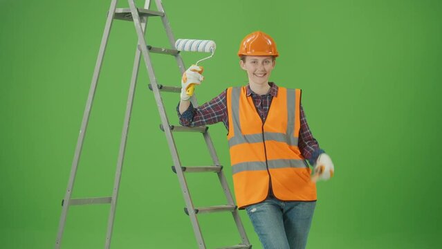 Green Screen.Young Happy Confident Female Construction Worker in Checkered Shirt,Safety Jacket,Helmet,Gloves,Leaning on a Ladder,Lifting the Paint Roller and Spatula Up, Smiling.Women at Work Concept
