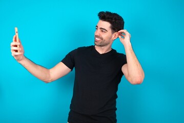 Young man wearing black T-shirt over blue studio background smiling and taking a selfie ready to...