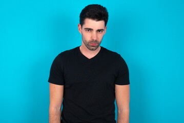 Obraz premium Offended dissatisfied Young man wearing black T-shirt over blue studio background with moody displeased expression at camera being disappointed by something