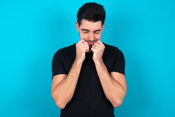 Pleased Young man wearing black T-shirt over blue studio background with closed eyes keeps hands near cheeks and smiles tenderly imagines something very pleasant