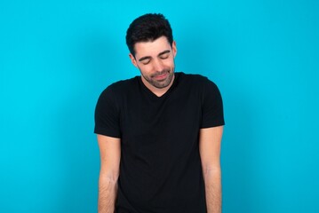 Obraz premium Positive Young man wearing black T-shirt over blue studio background with overjoyed expression closes eyes and laughs shows white perfect teeth