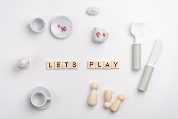 Flat lay of cute porcelain cups and saucers, fork, knife and peg dolls on white background. LETS PLAY written with tile letters