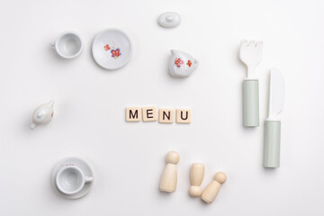 Flat lay of cute porcelain cups and saucers, fork, knife and peg dolls on white background. MENU written with tile letters