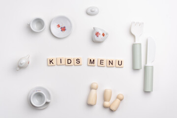 Flat lay of cute porcelain cups and saucers, fork, knife and peg dolls on white background. KIDS MENU written with tile letters