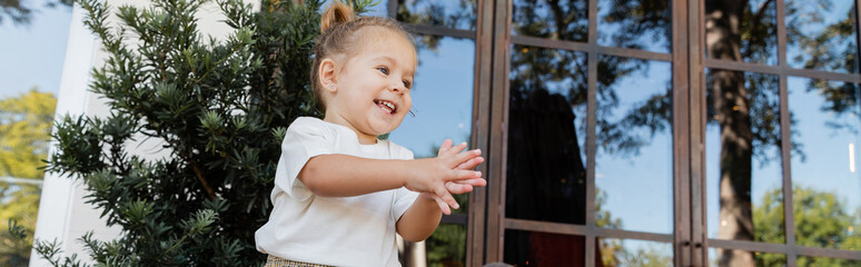 positive toddler girl in white t-shirt clapping hands near outdoor cafe in Miami, banner.