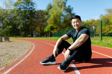 Young asian sportsman, spotter, runner sitting in stadium on treadmill and resting after jogging, exercise. Tired but smiling, he looks at the camera
