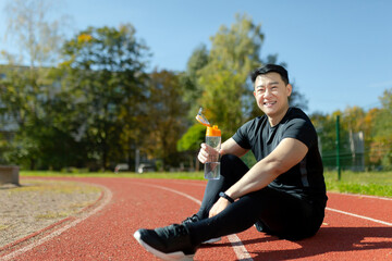 Young asian sportsman sitting in stadium on treadmill and resting after jogging, exercise. He holds...