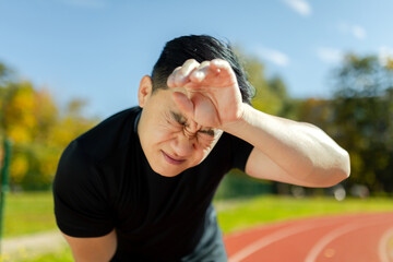 Close-up photo. Portrait of a young Asian man, an athlete standing in a stadium and holding his head. Feels severe pain, pressure, tension.