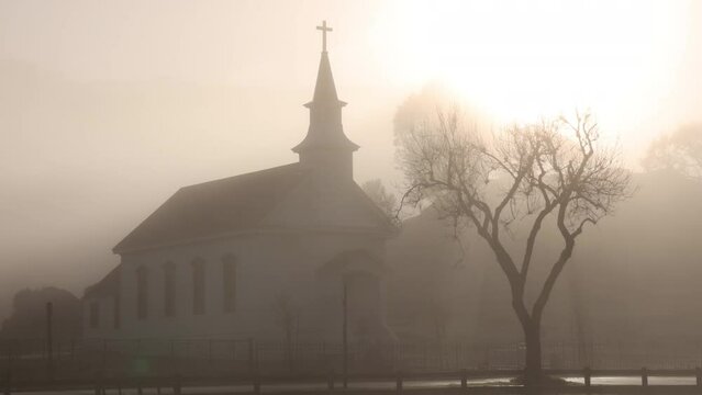 Classic car drives by small church in heavy fog with bright morning sun