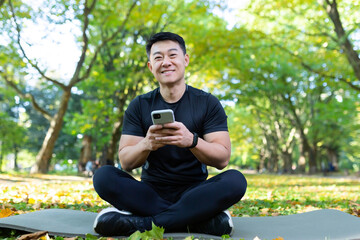 Portrait of a young Asian male athlete, coach sitting in the park on a mat in the lotus position and using the phone. He looks at the camera, smiles.