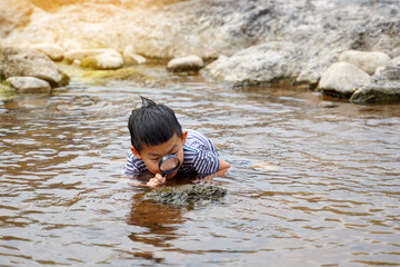 Asian boy enjoys exploring creatures in stream, along rocks with magnifying glass. concept of learning outside the classroom, natural learning resources. Soft and selective focus.