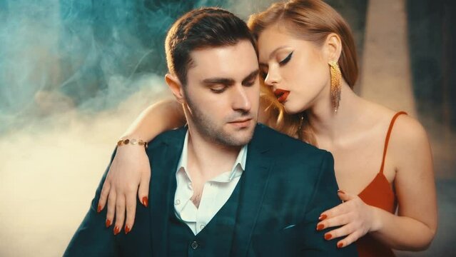 Sexy couple man and blonde woman in love. Red dress female hand touches strokes chest of stylish guy. Portrait Fashion model girl evening makeup red lips beauty face posing in dark room full smoke 4k