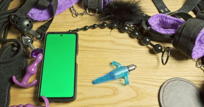 Small vibrator with rings around the edges spins in circle on table next to smartphone with green screen. Top view, close-up. Concept of online app, store, sex shop.