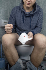 Hemorrhoid concept. A man sits on the toilet and holds white toilet paper in his hands. There is a little drop of blood on it. Aesthetic presentation of problems with the rectum.